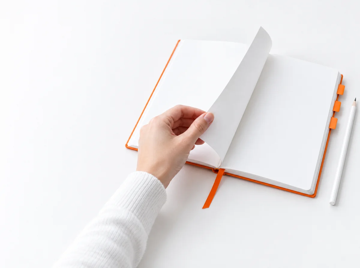 Illustration : une main tourne la page d'un carnet blanc à marque-page et onglets orange, un crayon posé à côté.